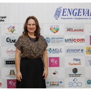 MULHER BRANCA, SORRINDO, CABELOS SOLTOS, CASTANHOS NA ALTURA DOS OMBROS, OLHOS CASTANHOS, VESTE BLUSA DE MANGA CURTA ESTAMPADA DE BEGE E PRETO, SAIA PRETA, COLAR PRETO. ESTÁ EM PÉ, DE FRENTE PARA A FOTO, BRAÇOS ESTICADOS AO LONGO DO CORPO. ENQUADRAMENTO J