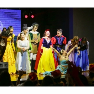 VÁRIAS MENINAS EM CIMA DO PALCO, CADA UMA REPRESENTANDO UMA PRINCESA DA DISNEY E TRÊS MENINOS VESTIDOS DE PRÍNCIPES. 